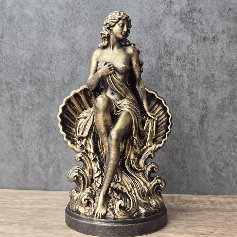 K&ouml;nnte beinhalten: Eine bronzefarbene Statue, die eine Figur zeigt, die in einer gro&szlig;en, geriffelten Muschel sitzt. Die Figur hat langes, welliges Haar und ist in flie&szlig;enden Stoff geh&uuml;llt, der den Unterk&ouml;rper bedeckt, w&auml;hrend der Oberk&ouml;rper teilweise frei bleibt.
