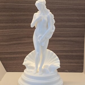 May include: A white matte sculpture of a standing figure with long flowing hair, positioned in front of a large open seashell. The figure is placed on a circular tiered base, capturing a classic form with smooth surfaces and detailed contours.