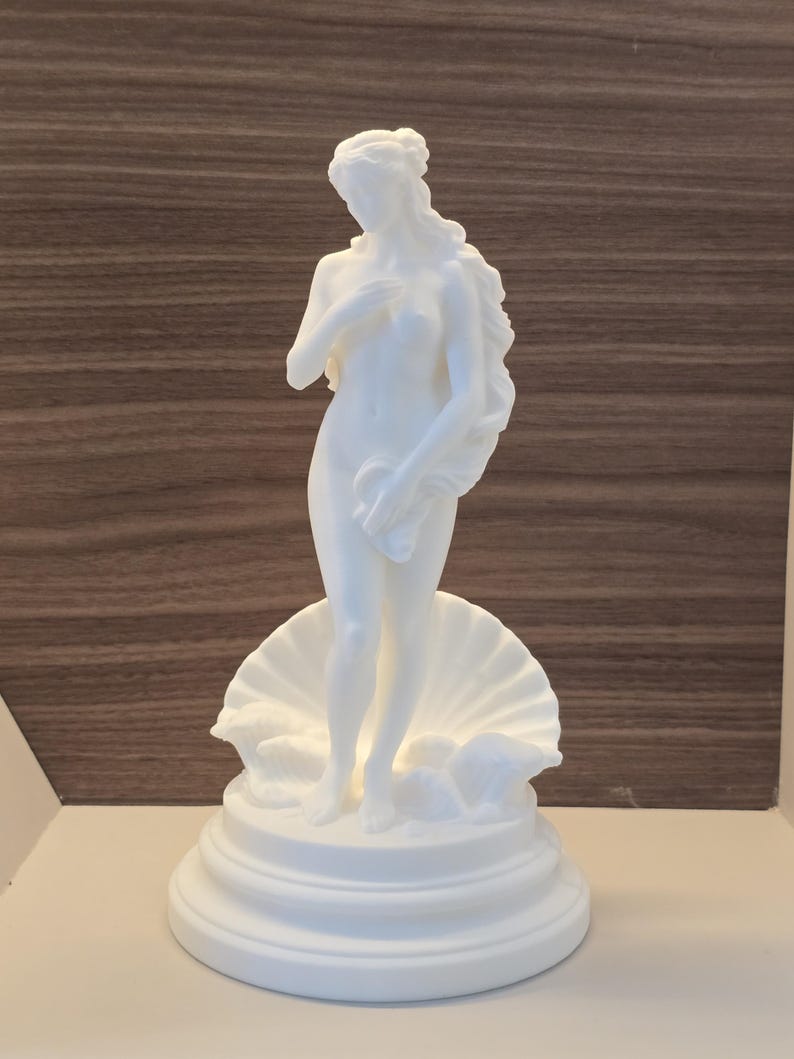 May include: A white matte sculpture of a standing figure with long flowing hair, positioned in front of a large open seashell. The figure is placed on a circular tiered base, capturing a classic form with smooth surfaces and detailed contours.