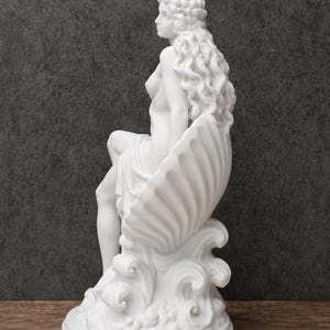 K&ouml;nnte beinhalten: Eine wei&szlig;e, matte Skulptur einer Figur mit langem, flie&szlig;endem, welligem Haar, die in einer gro&szlig;en, gerippten Muschel sitzt. Die Figur ist auf stilisierten, wirbelnden Wellen positioniert, die einen kreisf&ouml;rmigen Sockel bilden.