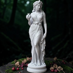 Op de afbeelding: Een wit, steenachtig standbeeld van een staande figuur in een gedrapeerd gewaad met een bloemenkrans. De figuur houdt een kleine granaatappel in de rechterhand, terwijl de linkerhand de zoom van het vloeiende, geplooide gewaad optilt.