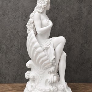 K&ouml;nnte beinhalten: Eine wei&szlig;e, matte Skulptur einer Figur mit langem, flie&szlig;endem Haar, die in einer gro&szlig;en Muschel sitzt, welche auf stilisierten Meereswellen ruht. Die Figur ist im Profil dargestellt, eine Hand an der Brust und Stoff &uuml;ber die Beine drapiert.