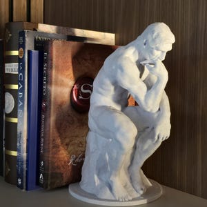 Könnte beinhalten: Eine weiße Skulptur eines sitzenden Mannes in nachdenklicher Pose, das Kinn auf der Hand. Die Skulptur steht auf einem runden Sockel, neben einem Stapel Bücher mit Titeln auf Spanisch und Englisch.