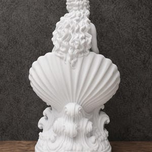 K&ouml;nnte beinhalten: Eine mattwei&szlig;e Skulptur, die eine Figur mit langem, flie&szlig;endem, welligem Haar zeigt, die in einer gro&szlig;en, vertikal gerippten Muschel sitzt. Der Sockel der Skulptur weist stilisierte, wirbelnde Wellenmuster auf, die ein einheitliches Design bilden.