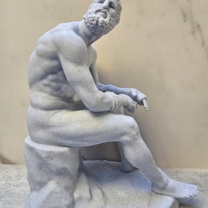 May include: A light blue statue of a seated man with a beard, resting on a rock-like base. The sculpture has detailed musculature and a pensive expression, with one leg extended and the other bent. The statue is approximately 12 inches tall.