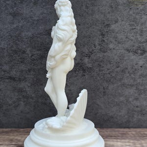 May include: A white matte sculpture of a standing figure with long flowing hair, positioned inside a large seashell. The figure is mounted on a circular tiered base, showcasing smooth surfaces and detailed contours throughout the entire form.
