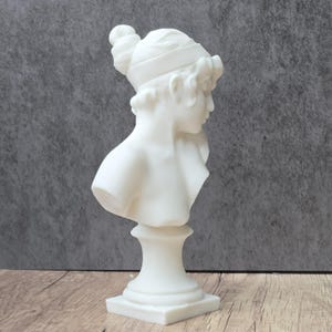 May include: A white matte bust sculpture depicting a profile view of a figure with hair styled in a bun and a headband. The sculpture features a smooth finish, a neck, and shoulders, resting on a circular pedestal base atop a square plinth.