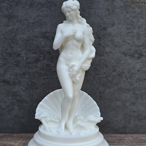 May include: A white matte sculpture of a standing figure with long flowing hair, partially covering the body with one hand while holding a draped cloth with the other. The figure stands atop a large seashell base with stylized waves.