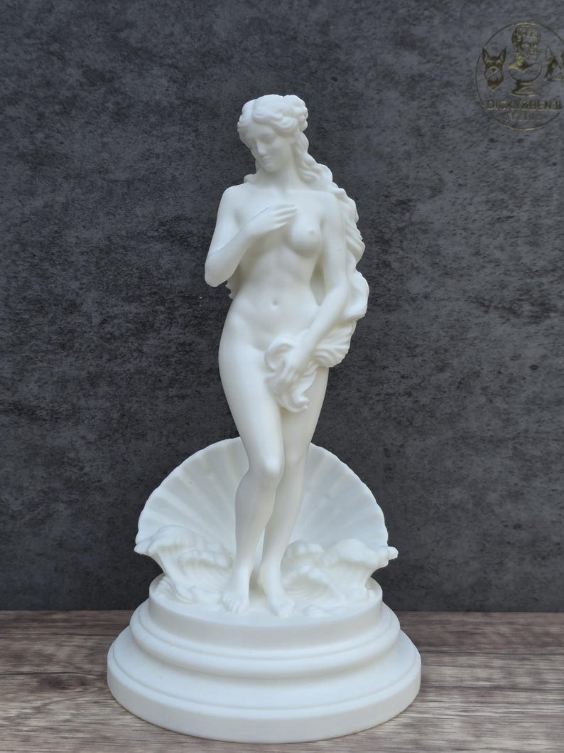 May include: A white matte sculpture of a standing figure with long flowing hair, partially covering the body with one hand while holding a draped cloth with the other. The figure stands atop a large seashell base with stylized waves.