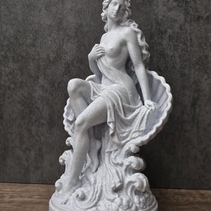K&ouml;nnte beinhalten: Eine wei&szlig;e, gesprenkelte Statue einer Figur mit langem, flie&szlig;endem Haar, die in einer gro&szlig;en, offenen Muschel sitzt. Die Figur ist teilweise mit Stoff drapiert und ruht auf einem Sockel mit skulptierten, wellenf&ouml;rmigen Details am Fu&szlig;.