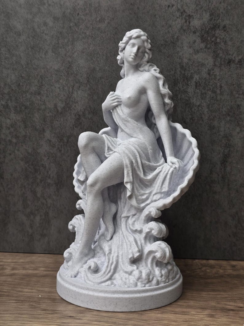 K&ouml;nnte beinhalten: Eine wei&szlig;e, gesprenkelte Statue einer Figur mit langem, flie&szlig;endem Haar, die in einer gro&szlig;en, offenen Muschel sitzt. Die Figur ist teilweise mit Stoff drapiert und ruht auf einem Sockel mit skulptierten, wellenf&ouml;rmigen Details am Fu&szlig;.