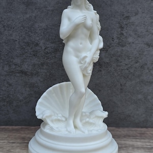 May include: A white matte sculpture of a standing figure with long flowing hair, partially covering the body with hands. The figure stands on a circular tiered base in front of a large scalloped seashell and stylized waves, all in a solid white finish.