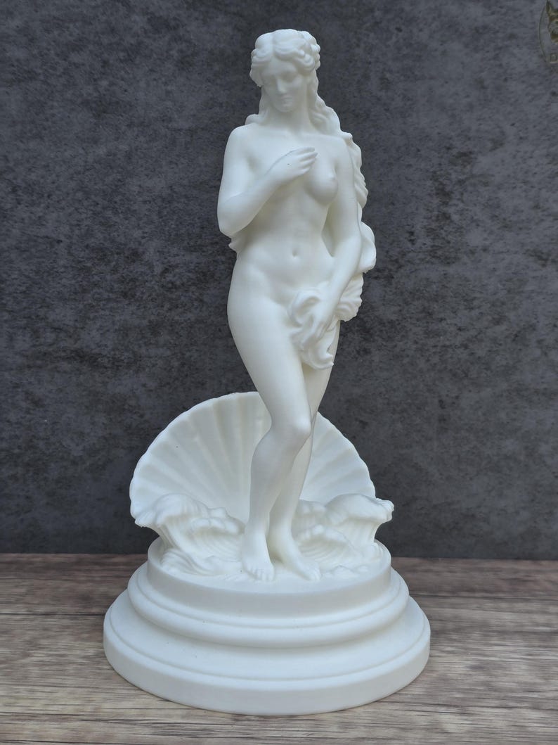 May include: A white matte sculpture of a standing figure with long flowing hair, partially covering the body with hands. The figure stands on a circular tiered base in front of a large scalloped seashell and stylized waves, all in a solid white finish.