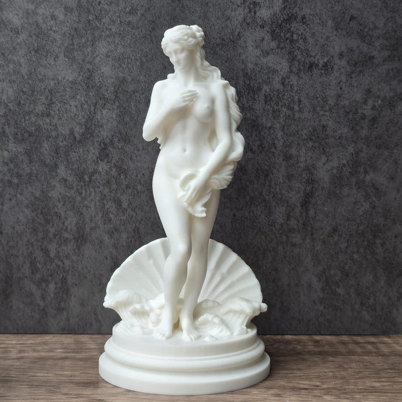 May include: A white matte sculpture of a standing figure with long hair, partially covered by a draped cloth, positioned in front of a large scallop shell base with stylized waves. The figure is depicted in a classic pose with one hand on the chest.
