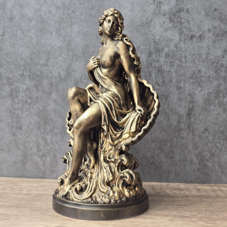 K&ouml;nnte beinhalten: Eine bronzefarbene Statue einer Figur, die in einer gro&szlig;en Muschel sitzt. Die Figur hat langes, flie&szlig;endes Haar und ist teilweise mit Stoff bedeckt, w&auml;hrend sie sich an die Muschelstruktur lehnt, die von wirbelnden Wellenelementen getragen wird.