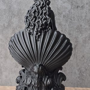 K&ouml;nnte beinhalten: Eine mattschwarze Skulptur, die eine Figur mit langem, flie&szlig;endem, welligem Haar zeigt, die in einer gro&szlig;en, gerippten Muschel sitzt. Der Sockel weist komplizierte, wirbelnde, organische Verzierungen auf, und das gesamte St&uuml;ck ist in einheitlichem Schwarz gehalten.