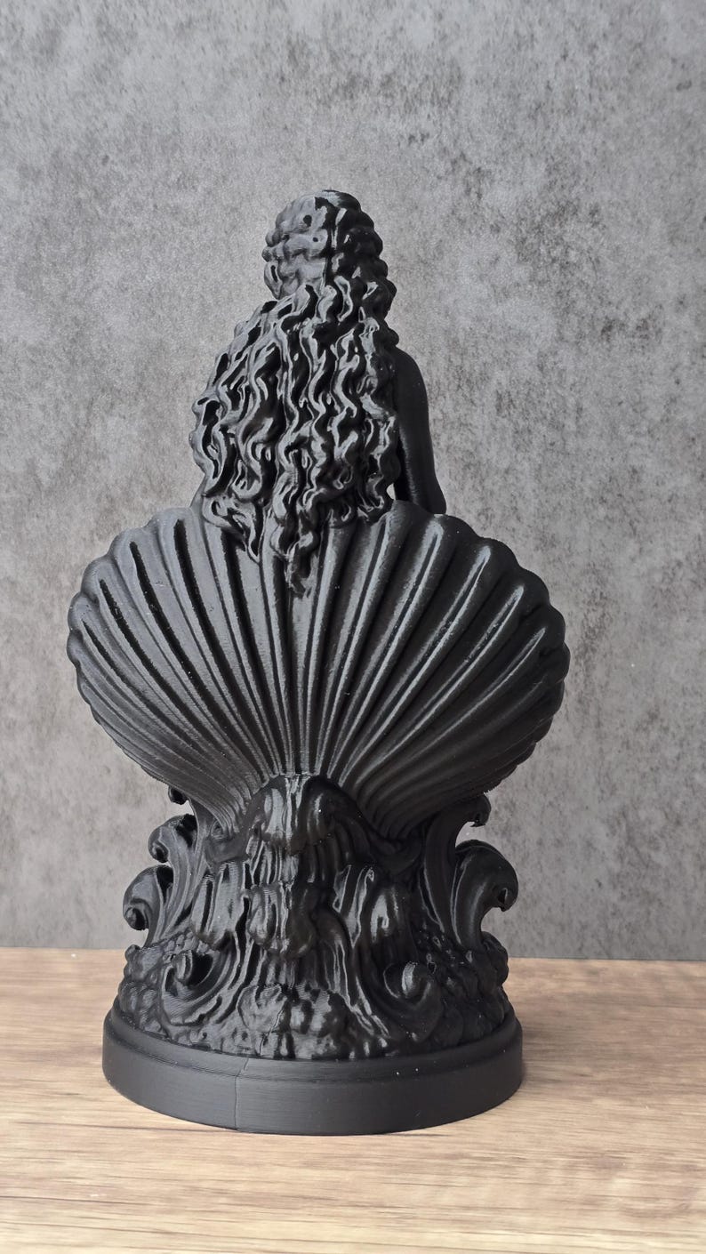 K&ouml;nnte beinhalten: Eine mattschwarze Skulptur, die eine Figur mit langem, flie&szlig;endem, welligem Haar zeigt, die in einer gro&szlig;en, gerippten Muschel sitzt. Der Sockel weist komplizierte, wirbelnde, organische Verzierungen auf, und das gesamte St&uuml;ck ist in einheitlichem Schwarz gehalten.