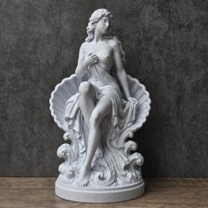K&ouml;nnte beinhalten: Eine wei&szlig;e, gesprenkelte steinerne Statue einer Figur, die in einer gro&szlig;en, offenen Muschel sitzt. Die Figur hat langes, flie&szlig;endes Haar, ist teilweise mit Stoff bedeckt und steht auf einem Sockel mit skulptierten, wellenf&ouml;rmigen Elementen.