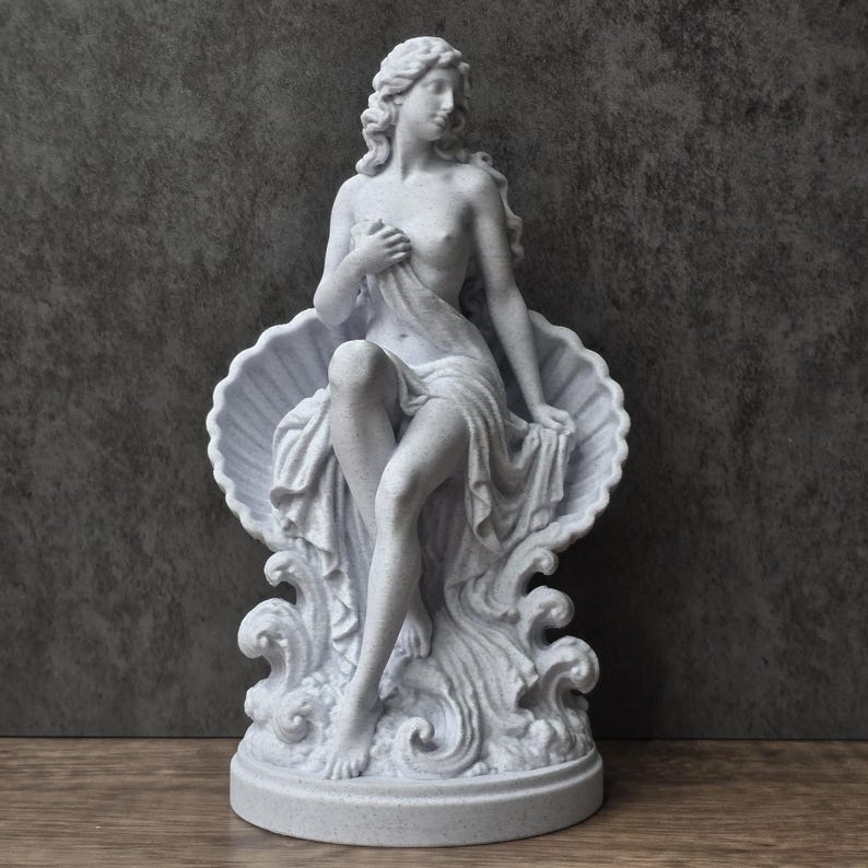 K&ouml;nnte beinhalten: Eine wei&szlig;e, gesprenkelte steinerne Statue einer Figur, die in einer gro&szlig;en, offenen Muschel sitzt. Die Figur hat langes, flie&szlig;endes Haar, ist teilweise mit Stoff bedeckt und steht auf einem Sockel mit skulptierten, wellenf&ouml;rmigen Elementen.