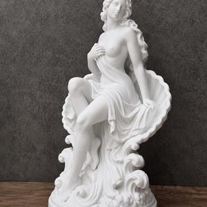 K&ouml;nnte beinhalten: Eine wei&szlig;e, matte Skulptur einer Figur mit langem, flie&szlig;endem Haar, die in einer gro&szlig;en, muschelf&ouml;rmigen Schale sitzt. Die Figur ist teilweise mit Stoff bedeckt, der &uuml;ber stilisierte Wellen f&auml;llt und auf einem runden Sockel ruht.