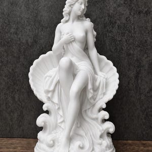 K&ouml;nnte beinhalten: Eine wei&szlig;e, matte Skulptur einer Figur mit langem, flie&szlig;endem Haar, die in einer gro&szlig;en, muschelf&ouml;rmigen Schale sitzt. Die Figur ist teilweise mit Stoff drapiert und ruht auf stilisierten Wellen auf einem runden Sockel.