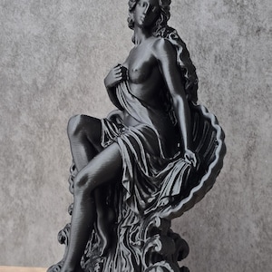 K&ouml;nnte beinhalten: Eine mattschwarze Figur, die eine sitzende Gestalt mit langem, flie&szlig;endem Haar darstellt, die teilweise in Stoff geh&uuml;llt ist und an einer gro&szlig;en, gewellten Muschel lehnt. Die Figur steht auf einer Basis aus stilisierten Wellen auf einem runden Sockel.