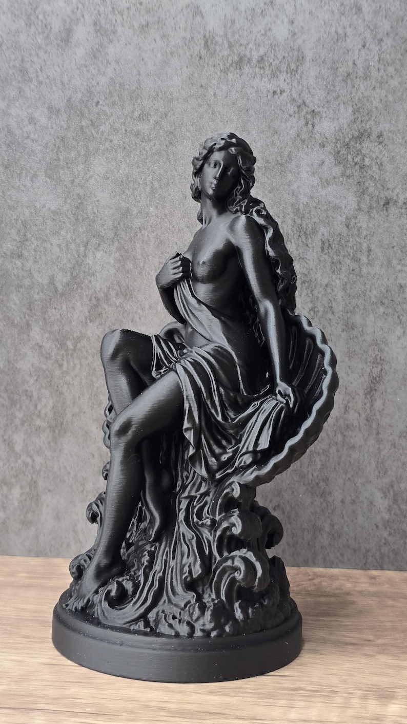 K&ouml;nnte beinhalten: Eine mattschwarze Figur, die eine sitzende Gestalt mit langem, flie&szlig;endem Haar darstellt, die teilweise in Stoff geh&uuml;llt ist und an einer gro&szlig;en, gewellten Muschel lehnt. Die Figur steht auf einer Basis aus stilisierten Wellen auf einem runden Sockel.