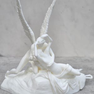 Puede incluir: Una escultura blanca que representa una escena clásica con dos figuras, una con grandes alas emplumadas, en un abrazo. Las figuras están sobre una base texturizada, con drapeado fluido. La escultura es de material opaco.