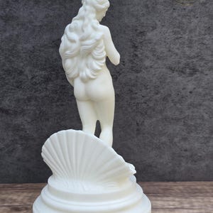 May include: A white statue of a figure with long flowing hair standing inside a large, ribbed seashell. The sculpture is positioned on a circular, tiered base. The figure is viewed from behind, showing detailed hair texture and a smooth finish.
