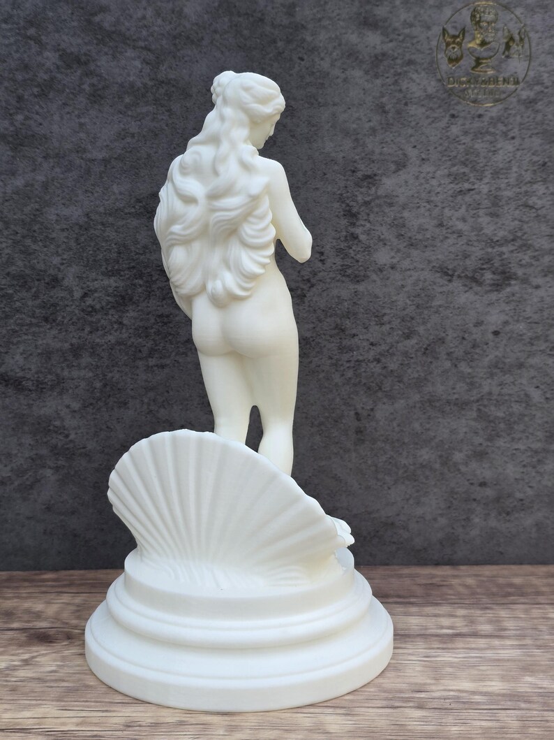 May include: A white statue of a figure with long flowing hair standing inside a large, ribbed seashell. The sculpture is positioned on a circular, tiered base. The figure is viewed from behind, showing detailed hair texture and a smooth finish.