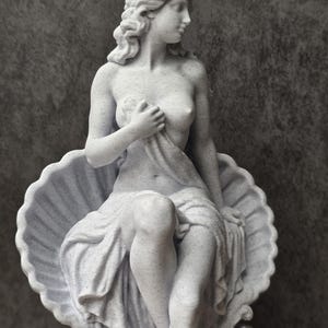 K&ouml;nnte beinhalten: Eine hellgraue, gesprenkelte Statue einer Figur mit welligem Haar, die in einer gro&szlig;en, geriffelten Muschel sitzt. Die Figur ist in Stoff geh&uuml;llt, der den Unterk&ouml;rper bedeckt und mit einer Hand an der Brust gehalten wird, w&auml;hrend sie nach rechts blickt.