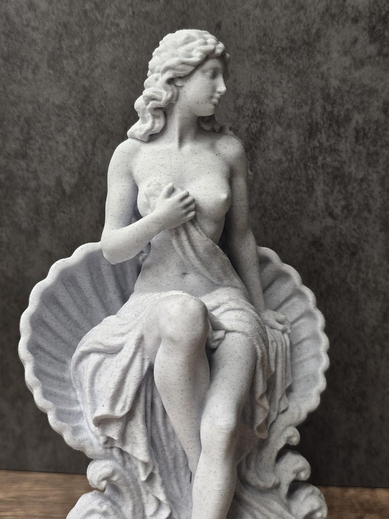 K&ouml;nnte beinhalten: Eine hellgraue, gesprenkelte Statue einer Figur mit welligem Haar, die in einer gro&szlig;en, geriffelten Muschel sitzt. Die Figur ist in Stoff geh&uuml;llt, der den Unterk&ouml;rper bedeckt und mit einer Hand an der Brust gehalten wird, w&auml;hrend sie nach rechts blickt.