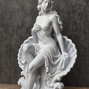 K&ouml;nnte beinhalten: Eine wei&szlig;e, gesprenkelte Statue mit matter Oberfl&auml;che, die eine Figur mit langem, flie&szlig;endem Haar zeigt, die in einer gro&szlig;en, offenen Jakobsmuschel sitzt. Die Figur ist teilweise mit Stoff bedeckt, eine Hand liegt auf der Brust.