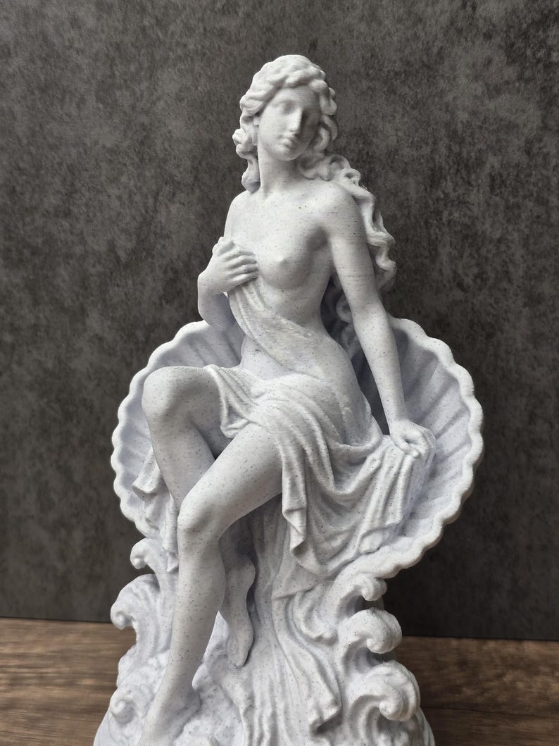 K&ouml;nnte beinhalten: Eine wei&szlig;e, gesprenkelte Statue mit matter Oberfl&auml;che, die eine Figur mit langem, flie&szlig;endem Haar zeigt, die in einer gro&szlig;en, offenen Jakobsmuschel sitzt. Die Figur ist teilweise mit Stoff bedeckt, eine Hand liegt auf der Brust.