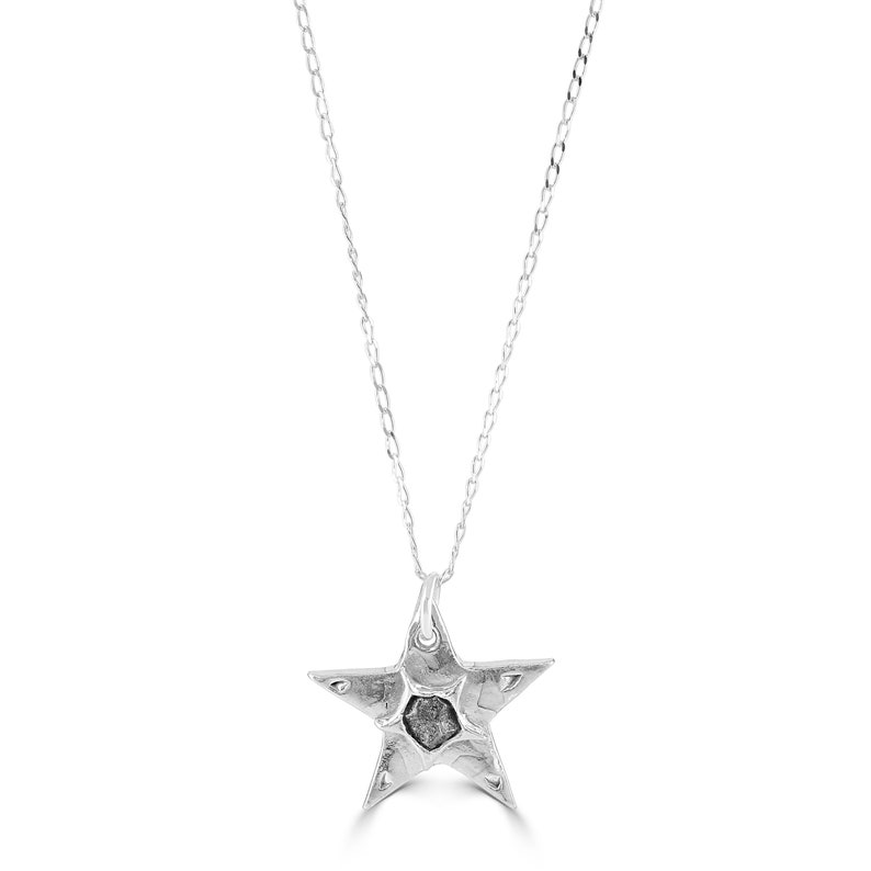 Puede incluir: Un collar de cadena de plata con un colgante de estrella de cinco puntas. La estrella tiene una superficie texturizada y una peque&ntilde;a piedra gris oscura en el centro.