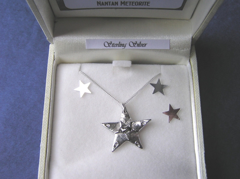 Puede incluir: Un conjunto de collar y pendientes de plata con un colgante de estrella de cinco puntas y pendientes a juego. El colgante es texturizado y tiene una forma ligeramente irregular. El conjunto se presenta en una caja blanca con las palabras "Nantan Meteorite" y "Sterling Silver" impresas en la tapa.