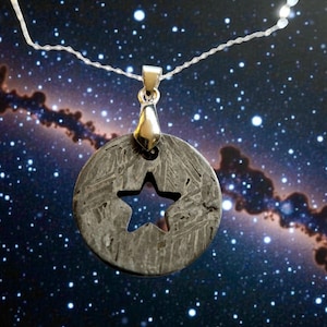 May include: A silver pendant necklace featuring a circular, gray stone pendant with a star-shaped cutout. The pendant hangs from a delicate silver chain. The background is a starry night sky.