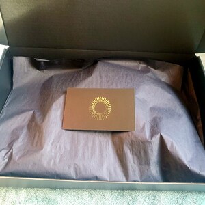 Puede incluir: Una caja negra con un forro de papel de seda negro. La caja est&aacute; abierta y se puede ver una peque&ntilde;a tarjeta marr&oacute;n con un dise&ntilde;o de c&iacute;rculo dorado en el interior.