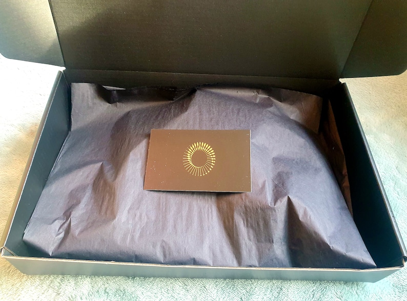 Puede incluir: Una caja negra con un forro de papel de seda negro. La caja est&aacute; abierta y se puede ver una peque&ntilde;a tarjeta marr&oacute;n con un dise&ntilde;o de c&iacute;rculo dorado en el interior.