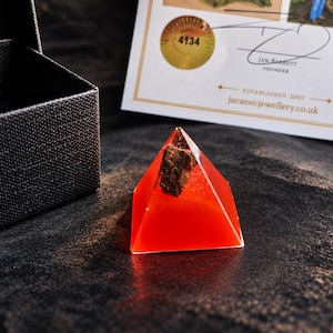 May include: A small, translucent, bright orange pyramid-shaped object made of resin, containing a dark, textured, rocky fragment embedded near the top point. The pyramid sits next to an open black box and a certificate of authenticity.