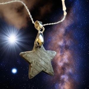 Può includere: Un ciondolo a forma di stella in materiale grigio strutturato è appeso a una catena d'argento. Il ciondolo è sospeso su uno sfondo di cielo stellato con una stella luminosa e radiosa.