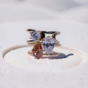 Puede incluir: Un anillo de varias bandas en tono dorado con tres piedras preciosas distintas. El diseño incluye una piedra redonda transparente, una piedra transparente en forma de pera y una piedra ovalada de color melocotón, montadas en una estructura metálica calada.