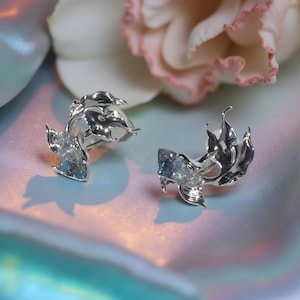 May include: A pair of silver-toned earrings with a fish-like design. Each earring features a stylized fish with flowing fins and a blue gemstone accent. The earrings are set against a soft, iridescent background.