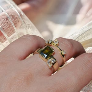 Puede incluir: Un anillo de oro con una gran piedra preciosa rectangular de color verde oliva flanqueada por dos pequeñas piedras claras en forma de pera, junto a una segunda banda de oro adornada con tres pequeñas piedras claras redondas en el borde.