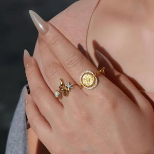 May include: Two gold rings with nautical themes. One ring features a compass design with a central starburst and surrounding clear stones. The other ring has a seashell, starfish, and fleur-de-lis design. Both rings are on a hand.