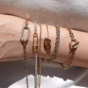 May include: An assortment of gold bracelets adorn a wrist. The bracelets include a chain link style with a crystal-covered oval, a safety pin design, a double heart, a tennis bracelet, and a curb chain with a crystal-accented toggle clasp. Jewelry.