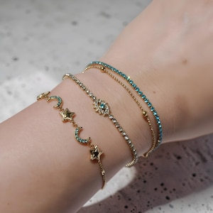 May include: A collection of gold-toned bracelets. One bracelet features a line of small, round, blue gemstones. Another has a series of crescent moons and stars with blue gemstones. The third bracelet has a floral design with a blue gemstone in the center.