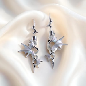 Puede incluir: Un par de pendientes plateados con temática de fantasía. Cada pendiente presenta un diseño de ala de dragón con acentos blancos iridiscentes. Los pendientes tienen un diseño artístico único con múltiples elementos decorativos.