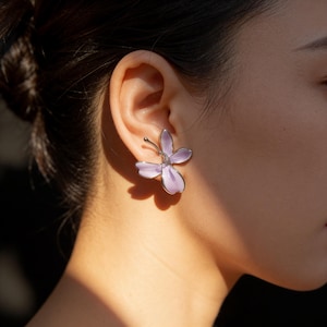 May include: A close-up of a lavender-colored flower-shaped earring. The earring has a silver-colored stem and a small, clear gem in the center. The earring is worn on the ear of a person with dark hair.