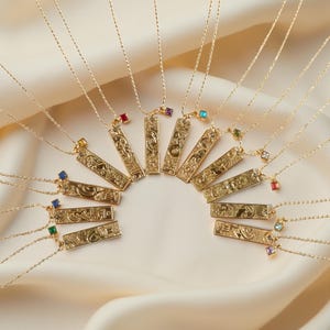 May include: A collection of gold-colored pendant necklaces arranged in a semi-circle. Each rectangular pendant features an engraved design and a small, colorful gemstone. The necklaces are displayed on a cream-colored fabric.